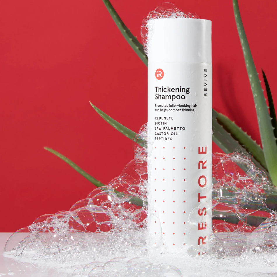 Image showing the Thickening Shampoo placed on top of a table with bubbles all over the bottle and showing an aloe vera behind the bottle.
