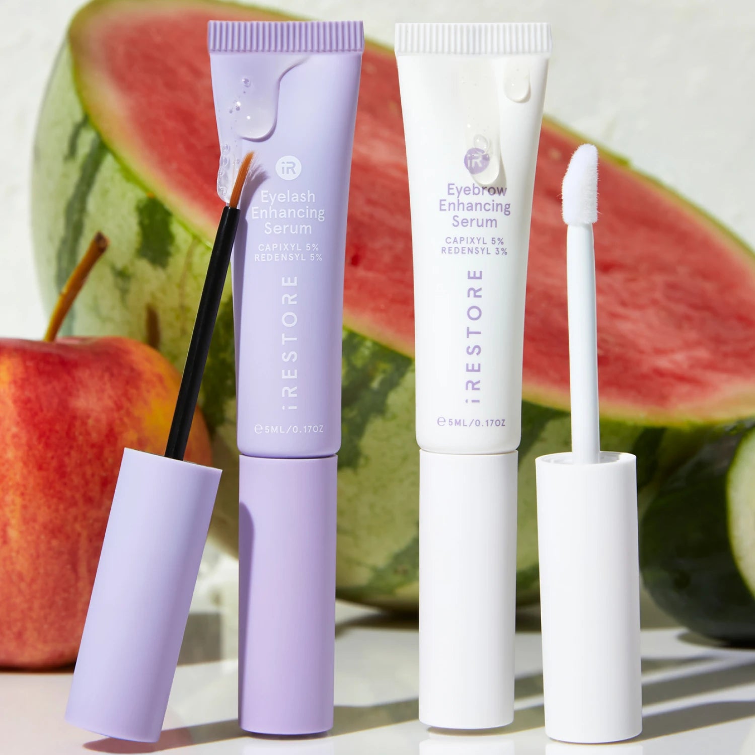 Image of the Eyebrow and Eyelash Enhancing Serum with the applicator beside each other showing apple and watermelon in the background.