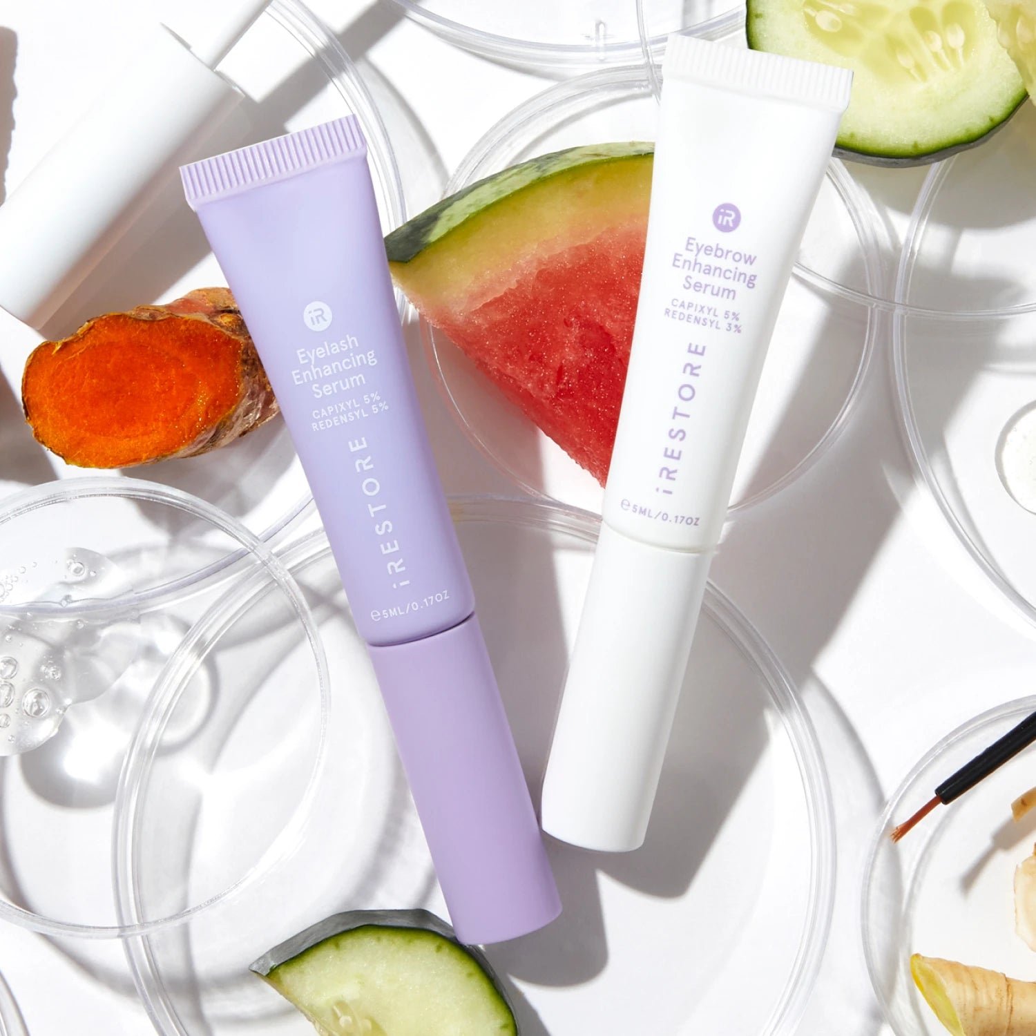 Lifestyle image of the Eyebrow and Eyelash Enhancing Serum beside each other with cucumber and watermelon in the background.