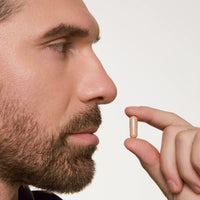A close-up side profile of a bearded man holding an Advanced Hair Growth Formula capsule pill between his fingers against a plain light background.