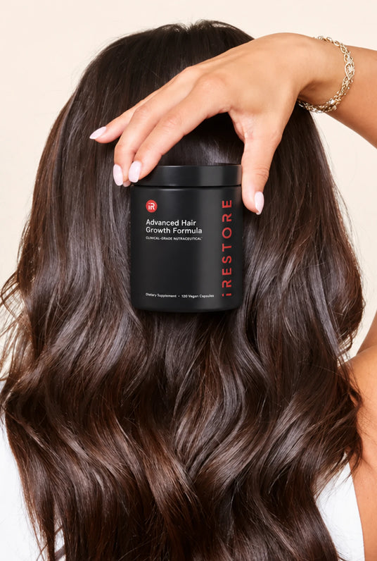 A woman with long, wavy dark brown hair holds a black jar of iRestore Advanced Hair Growth Formula behind her head. The background is a solid, warm off-white studio wall.
