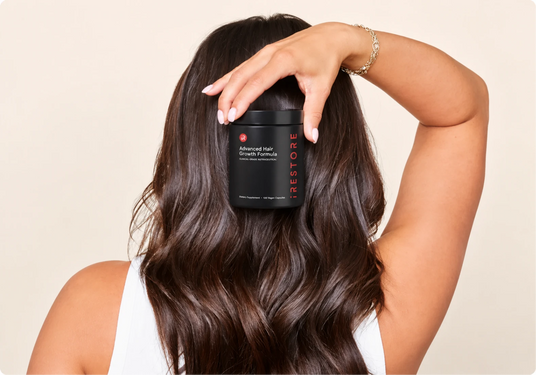 A woman with long, wavy dark brown hair holds a black jar of iRestore Advanced Hair Growth Formula behind her head. The background is a solid, warm off-white studio wall.