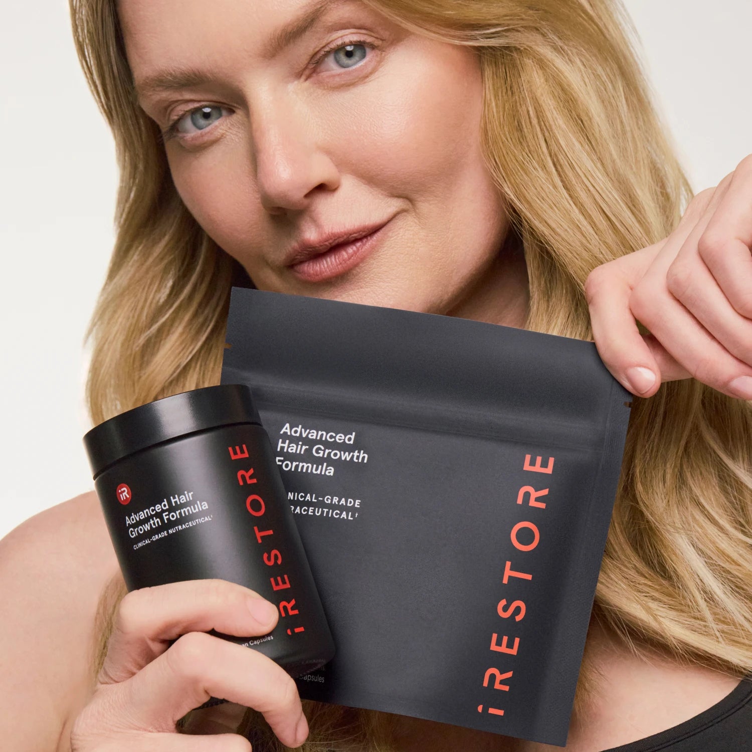 A woman holds a black jar and a black pouch of iRestore Advanced Hair Growth Formula against a clean white background.