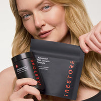 A woman holds a black jar and a black pouch of iRestore Advanced Hair Growth Formula against a clean white background.