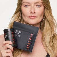 A woman holds a black jar and a black pouch of iRestore Advanced Hair Growth Formula against a clean white background.