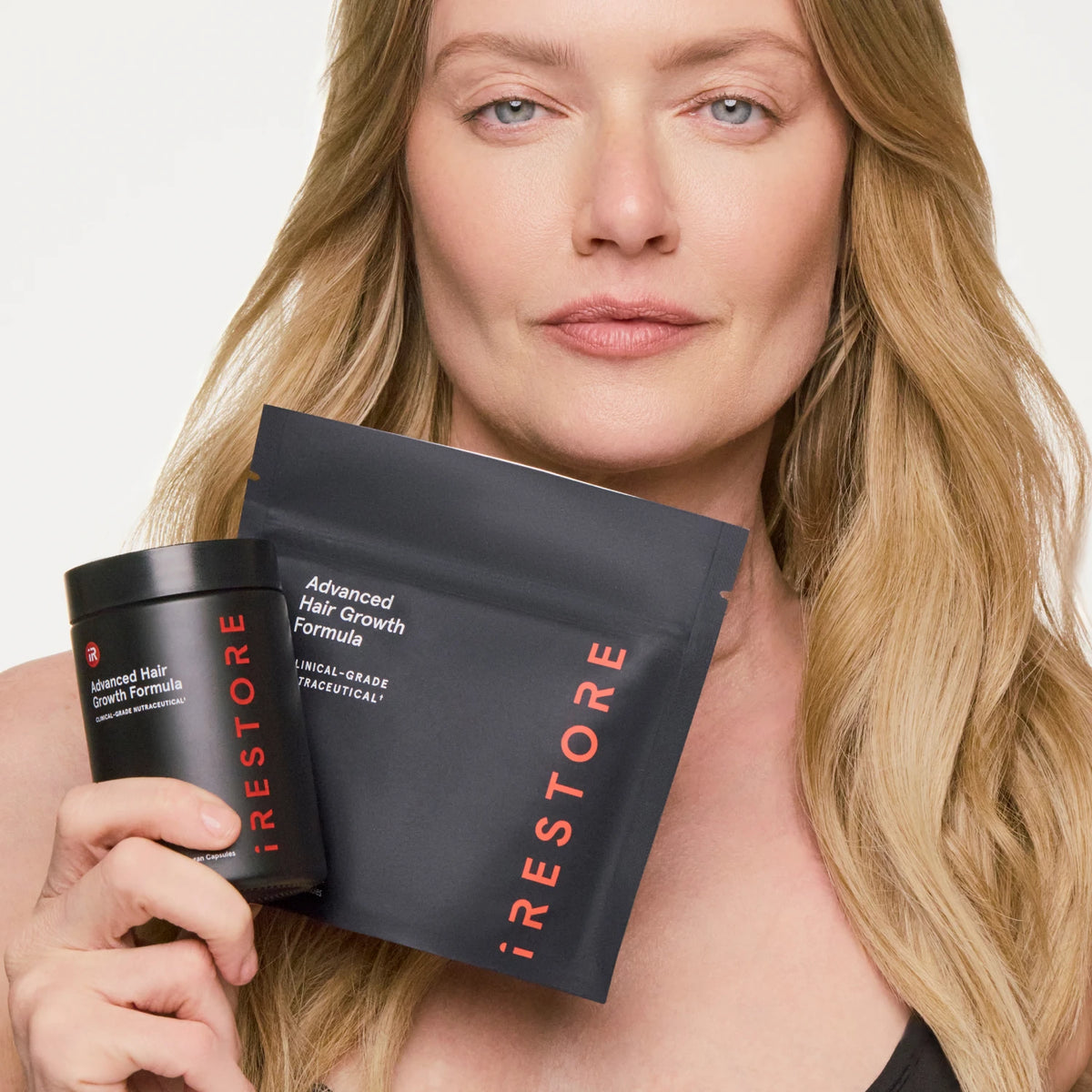 A woman holds a black jar and a black pouch of iRestore Advanced Hair Growth Formula against a clean white background.
