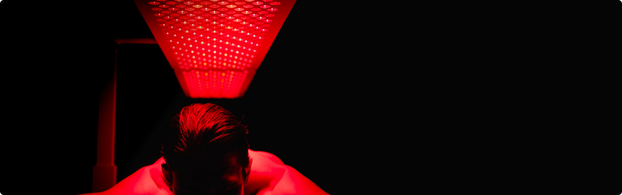  A close-up shot of a person lying down under a glowing red LED therapy panel in a dark room. The red light intensely illuminates the person's head and shoulders.