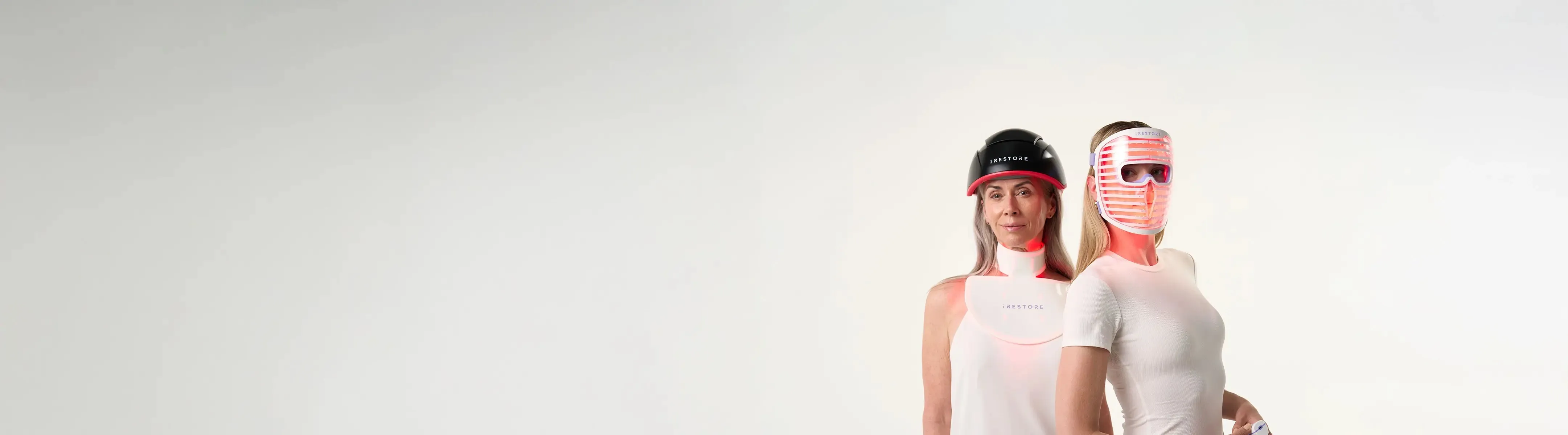 Mature woman wearing the Elite Hair Device standing beside a middle-aged woman wearing the Illumina LED Face Mask