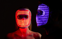 Front view of a woman wearing the iRESTORE LED face mask glowing red, standing beside a man in side view wearing the iRESTORE LED face mask glowing blue.