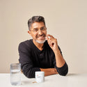 Image of a sitting man with Hair Growth Formula tablet in his hand. In front of him is a table with the cylindrical jar of the supplement placed on top of the table with a glass of water beside it.