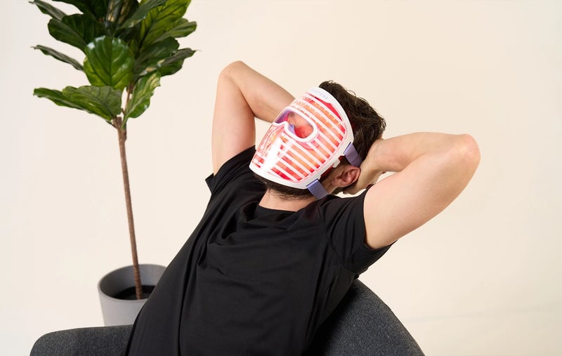 Man sitting and relaxing on a chair while wearing the iRESTORE LED face mask with red light turned on