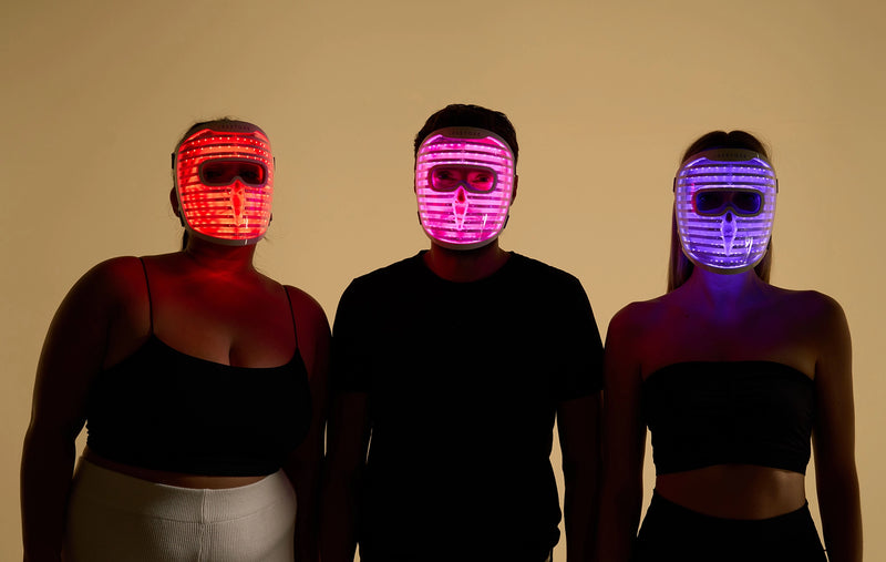 Man standing between two women, each wearing the iRESTORE LED face mask with the mask glowing in red, purple, and blue light, showcasing the different light therapy modes.