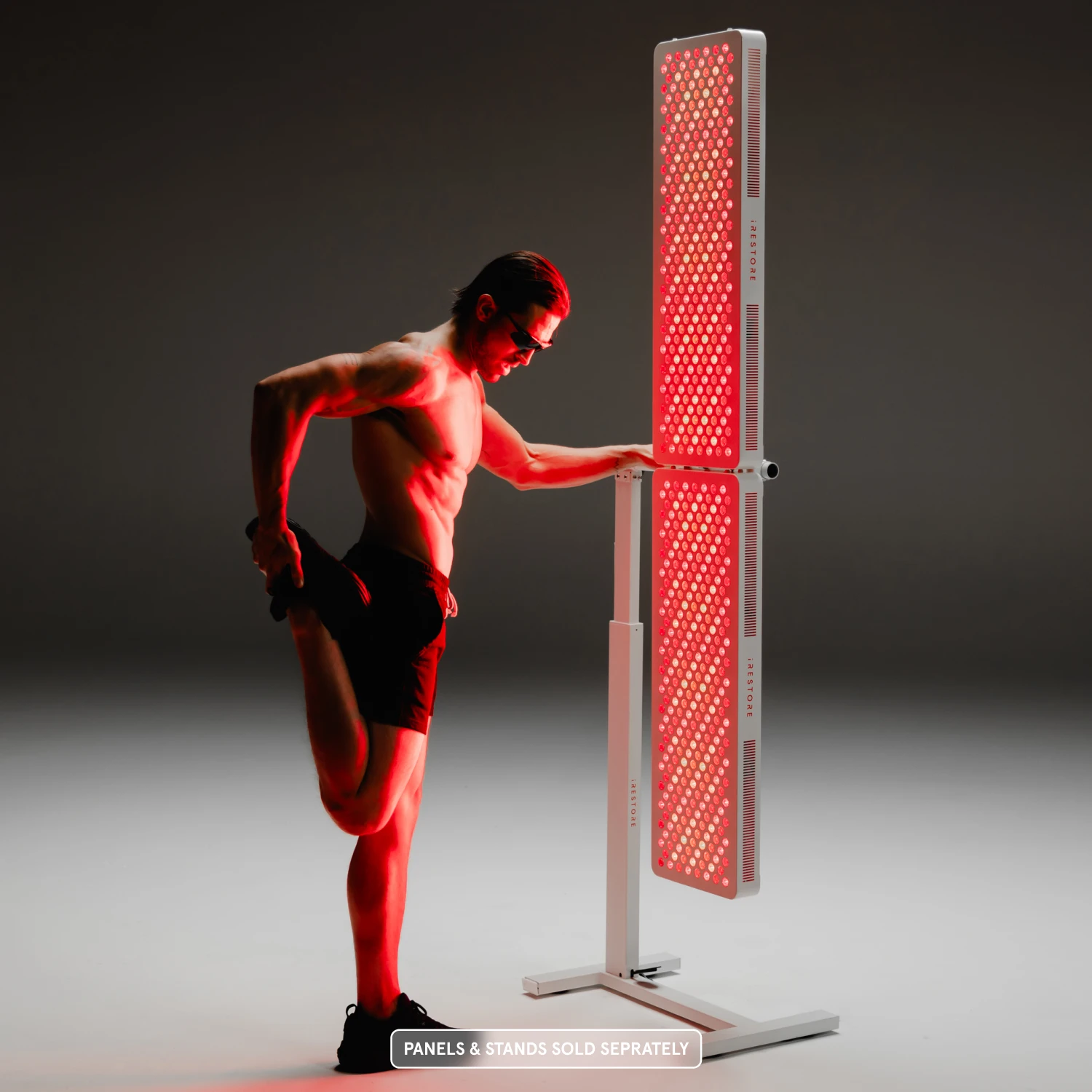 man in shorts and sunglasses performs a standing quad stretch while leaning on a tall, dual-panel red light therapy stand
