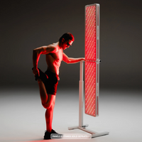 man in shorts and sunglasses performs a standing quad stretch while leaning on a tall, dual-panel red light therapy stand