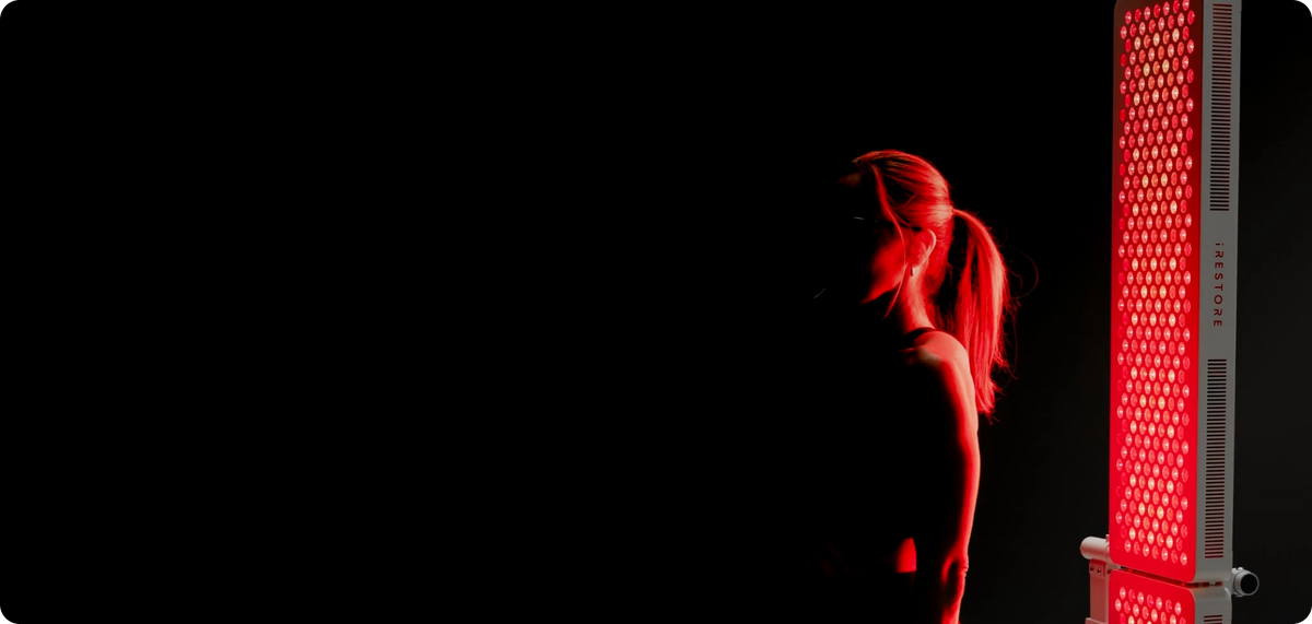 A person sits in a dark room, facing a large, glowing red light therapy panel that illuminates their face and torso.