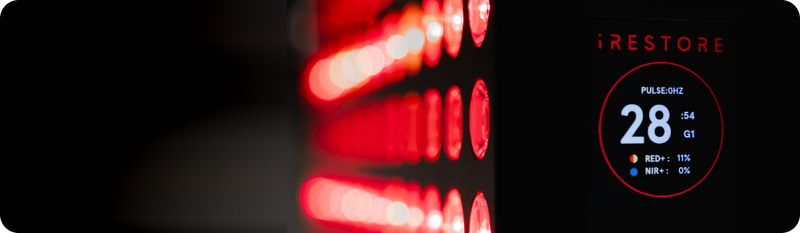 A close-up side view of an iRESTORE red light therapy device featuring a grid of glowing red LED lights and a digital display screen showing a timer