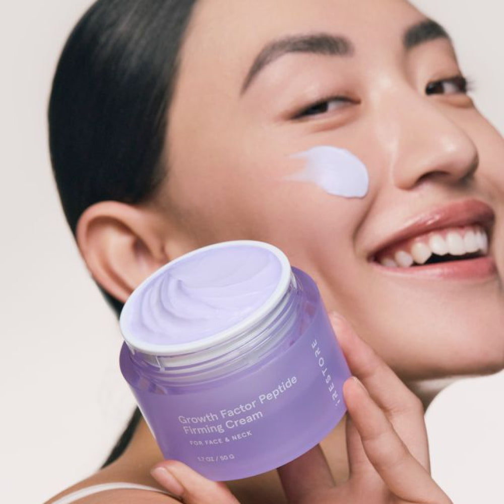 Smiling woman holding a jar of iRESTORE Growth Factor Peptide Firming Cream with a smudge of cream on her cheek