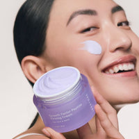 Smiling woman holding a jar of iRESTORE Growth Factor Peptide Firming Cream with a smudge of cream on her cheek