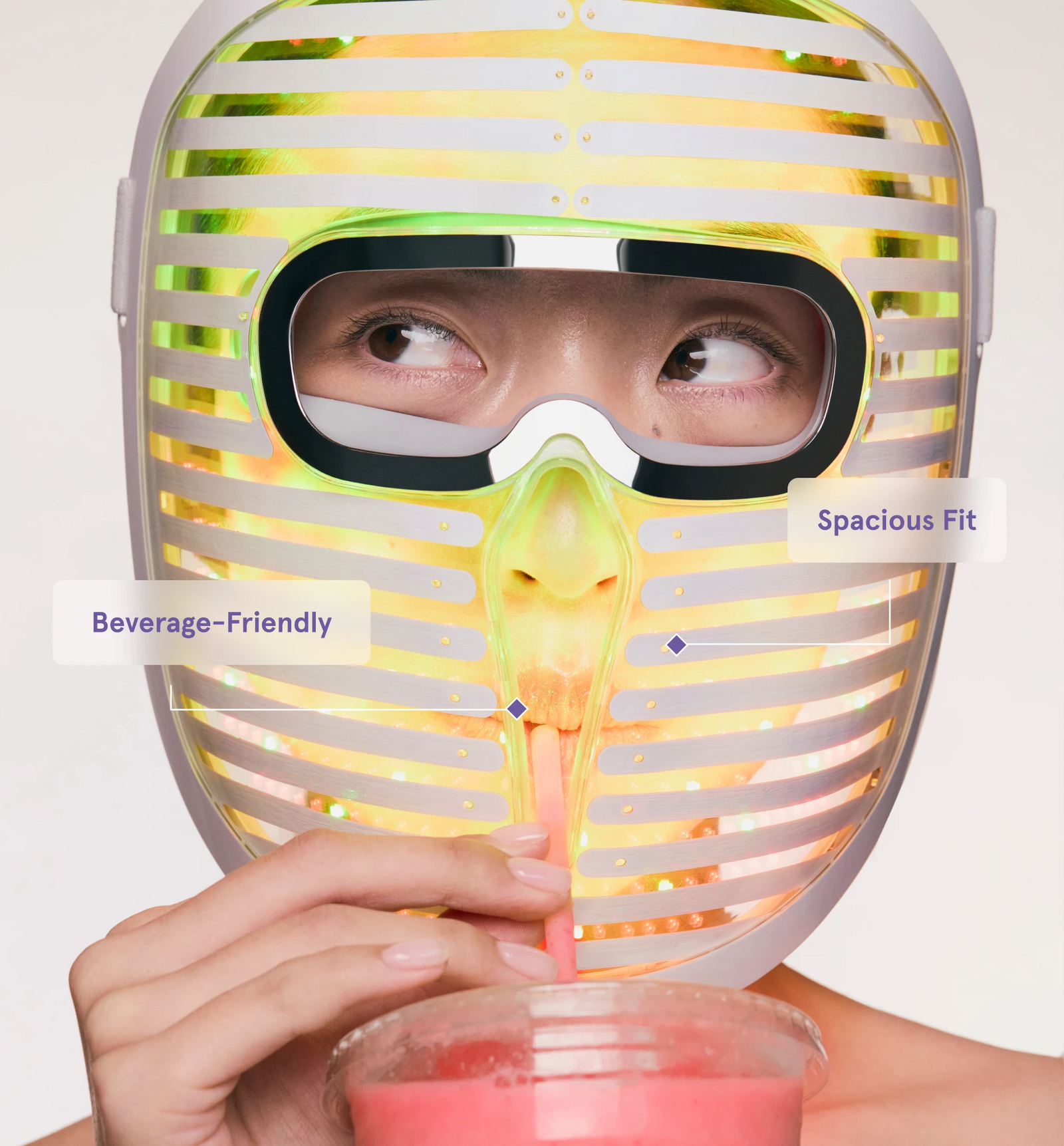 person wearing the Face Mask Pro glowing with yellow light while drinking through a straw, highlighting text labels for "Better Coverage" and "Spacious Fit."