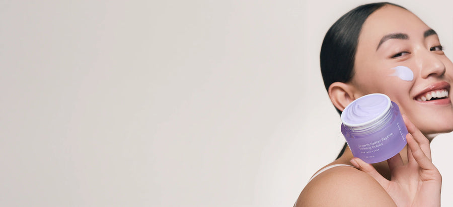 Smiling woman holding a jar of iRESTORE Growth Factor Peptide Firming Cream with a smudge of cream on her cheek