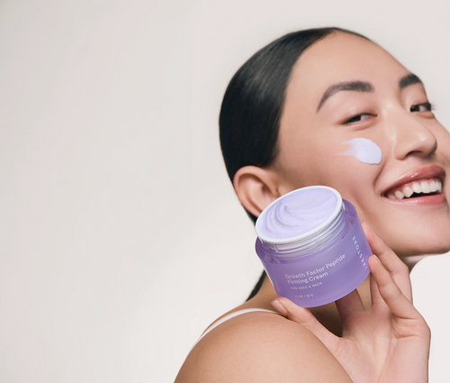 Smiling woman holding a jar of iRESTORE Growth Factor Peptide Firming Cream with a smudge of cream on her cheek