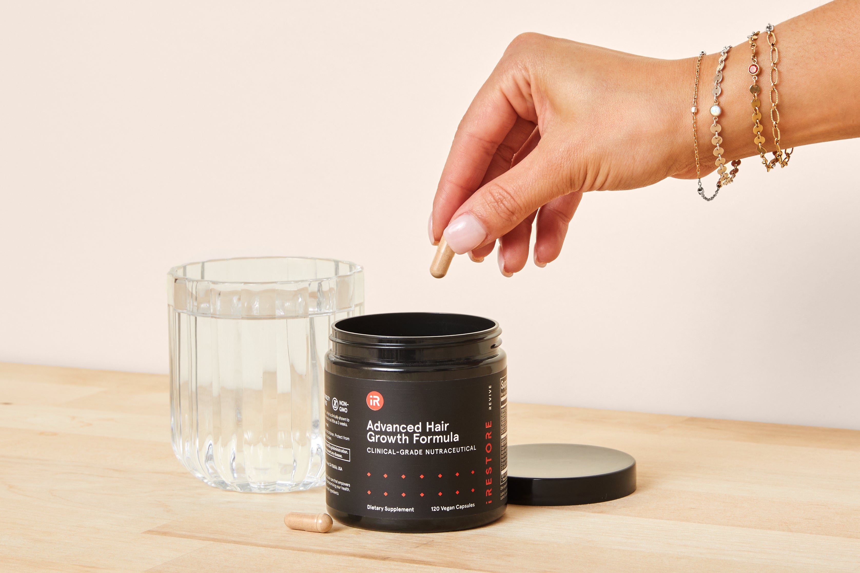 Hand taking a capsule from a jar labeled 'Advanced Hair Growth Formula' beside a glass of water.