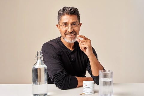 Image of a sitting man with Hair Growth Formula tablet in his hand. In front of him is a table with the cylindrical jar of the supplement placed on top of the table with a glass of water beside it.