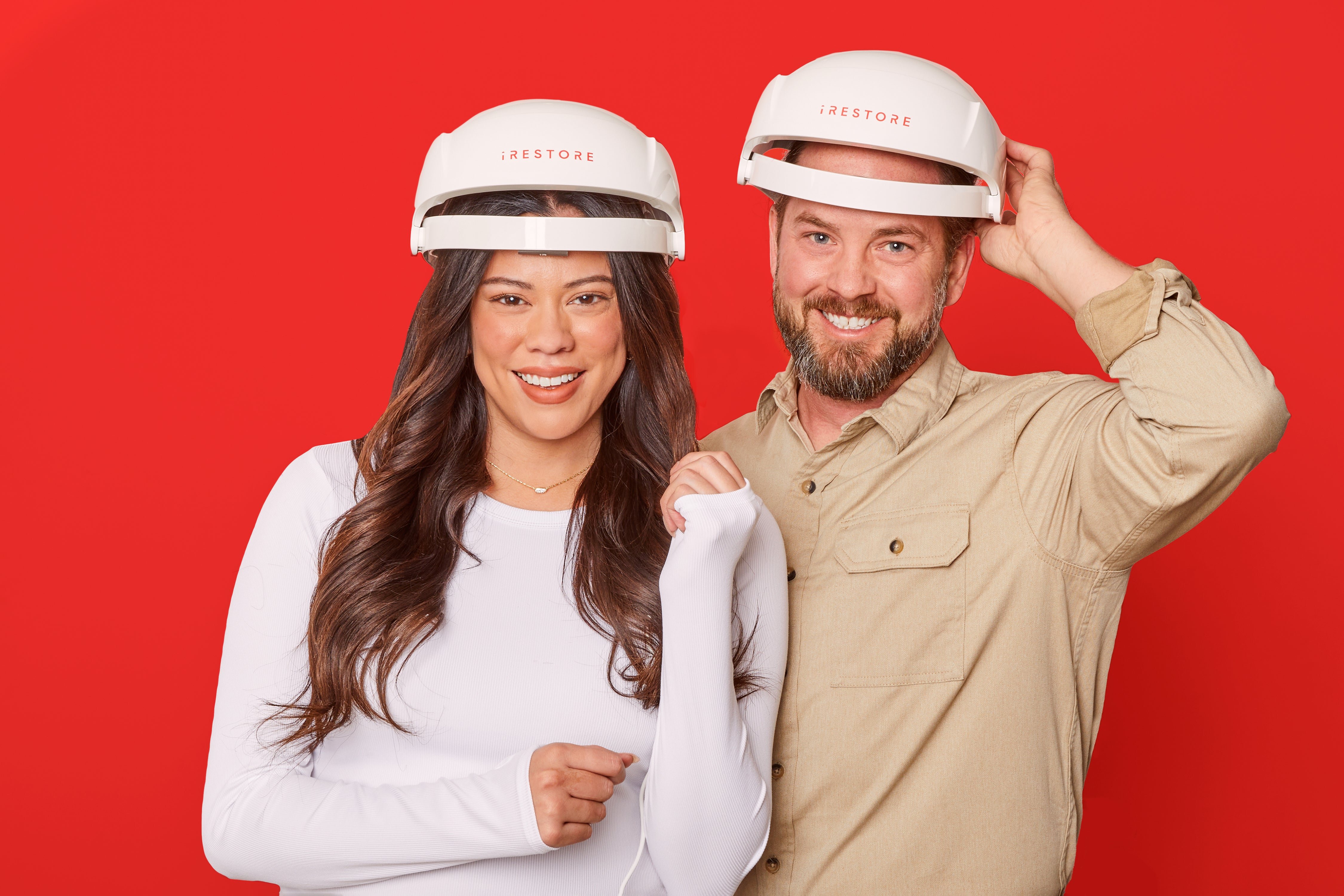 A woman and a man wearing white 'iRestore' helmets smile against a red background.