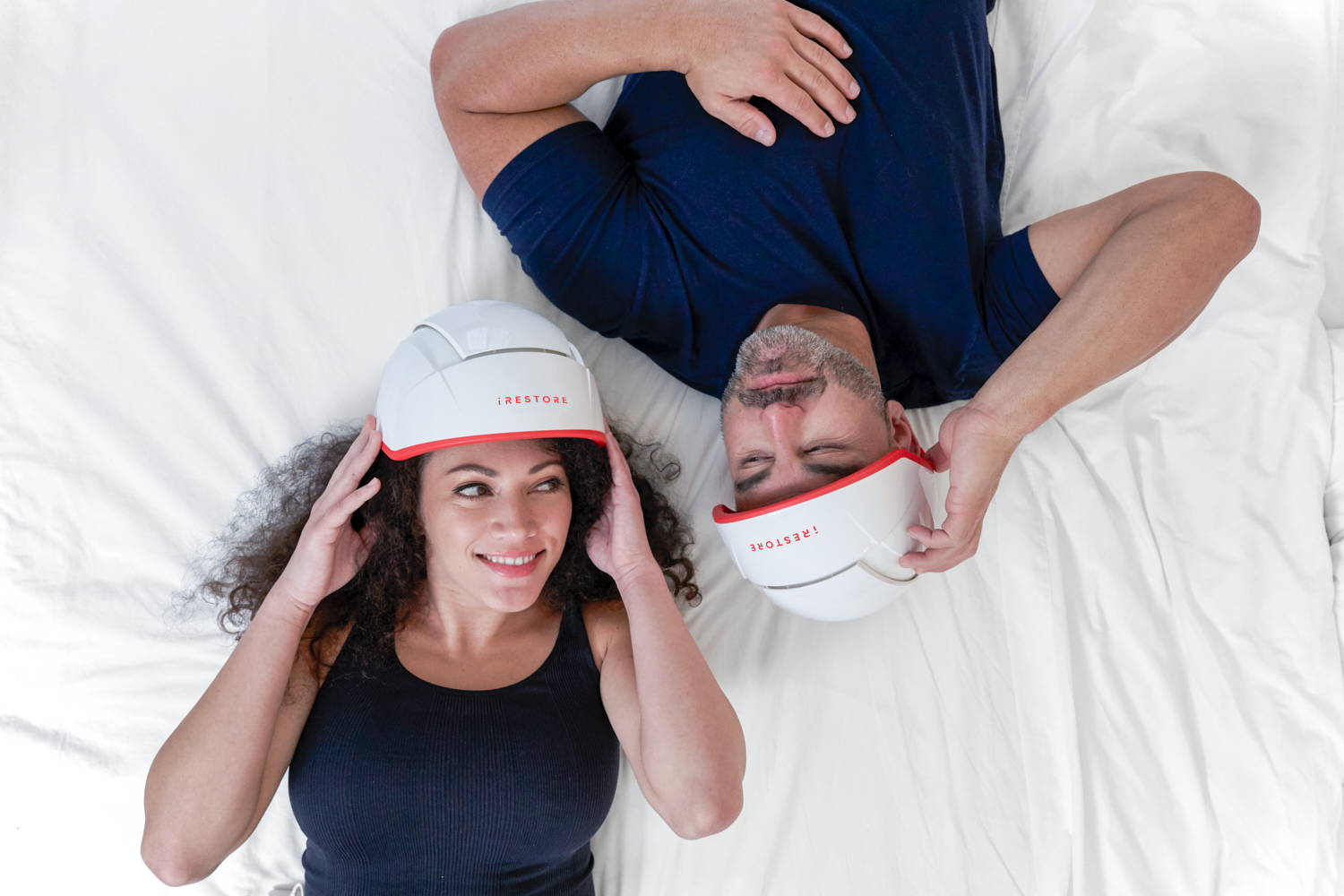 Two people lying down wearing iRestore hair growth helmets, smiling and looking relaxed.